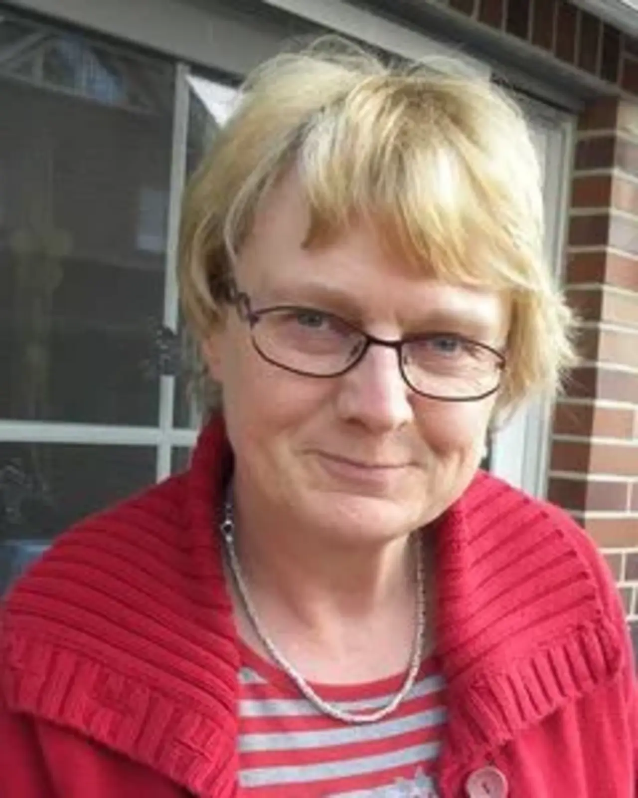 Porträtfoto einer Frau mit kurzem, blondem Haar und Brille. Sie trägt einen roten Pullover mit Kragen und ein gestreiftes T-Shirt. Der Hintergrund zeigt eine Hausfassade mit Fenstern.