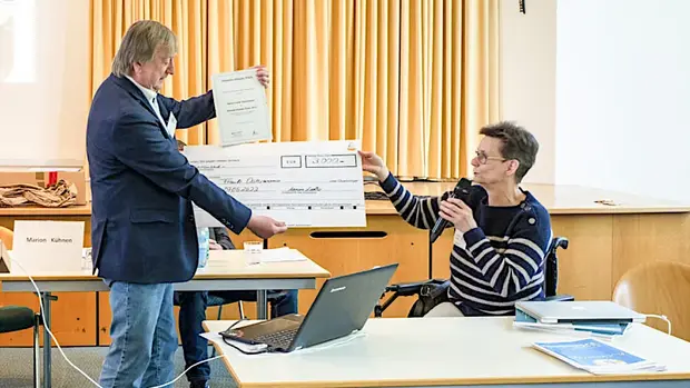 Foto von einer Preisverleihung in einem Veranstaltungsraum. Eine Frau im Rollstuhl nimmt einen symbolischen Scheck über 3.000 Euro von einem Mann entgegen, der zusätzlich eine Urkunde in der Hand hält. Die Frau spricht in ein Mikrofon. Im Hintergrund sind ein Vorhang und ein Konferenztisch mit Namensschildern zu sehen.