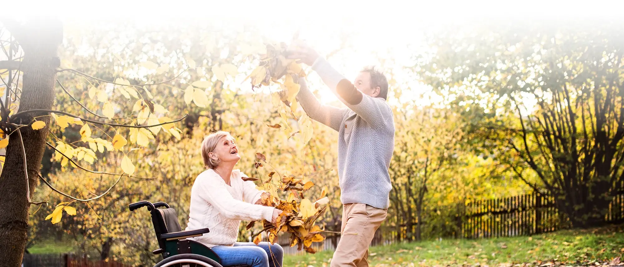 Seniorenpärchen im Herbstlaub. Sie sitzt im Rollstuhl. Er wirft Blätter über ihr in die Luft.