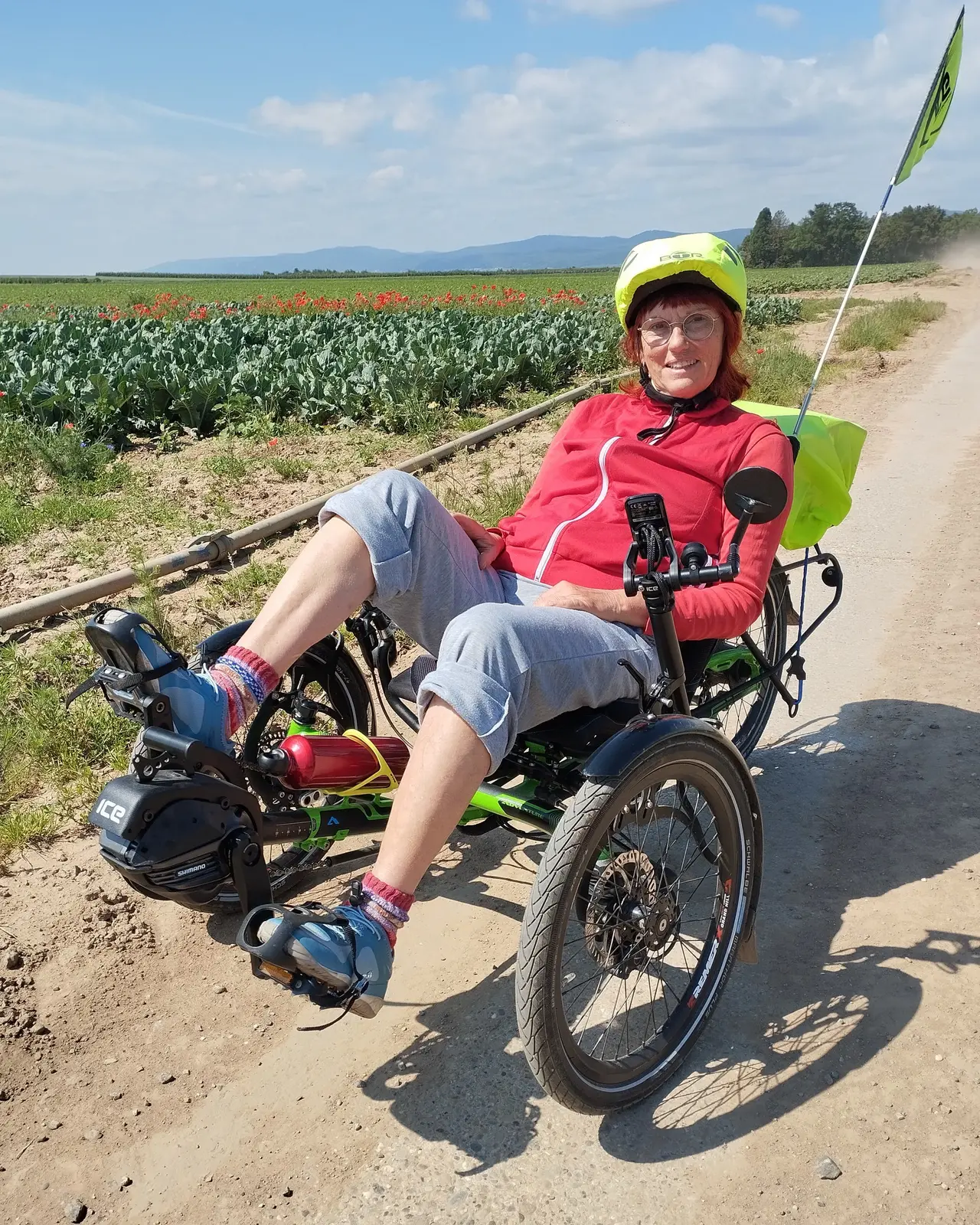 Eine Frau mittleren Alters sitzt entspannt auf einem Liegedreirad auf einem Feldweg. Sie trägt einen roten Pullover, eine graue Dreiviertelhose und eine gelbe Fahrradhelmhaube. Im Hintergrund sind Felder mit blühenden Pflanzen und eine hügelige Landschaft zu sehen. Sie lächelt in die Kamera und hält die Hände locker auf dem Lenker des Fahrrads.