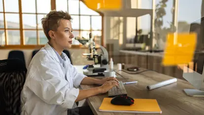 Eine Wissenschaftlerin mit kurzem Haar sitzt vor einem Computer und arbeitet konzentriert. Sie trägt einen weißen Kittel, und hinter ihr stehen ein Mikroskop und andere Laborgeräte.