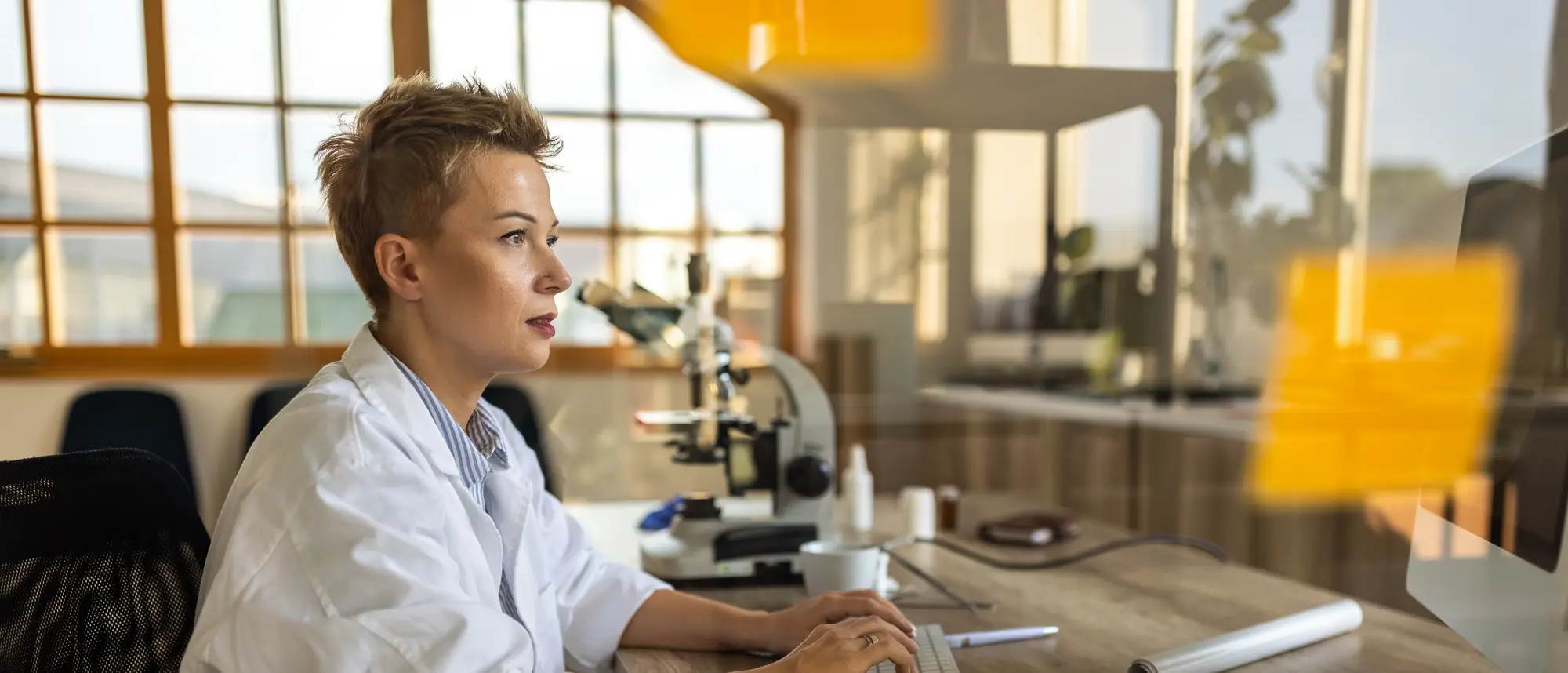 Eine Wissenschaftlerin mit kurzem Haar sitzt vor einem Computer und arbeitet konzentriert. Sie trägt einen weißen Kittel, und hinter ihr stehen ein Mikroskop und andere Laborgeräte.