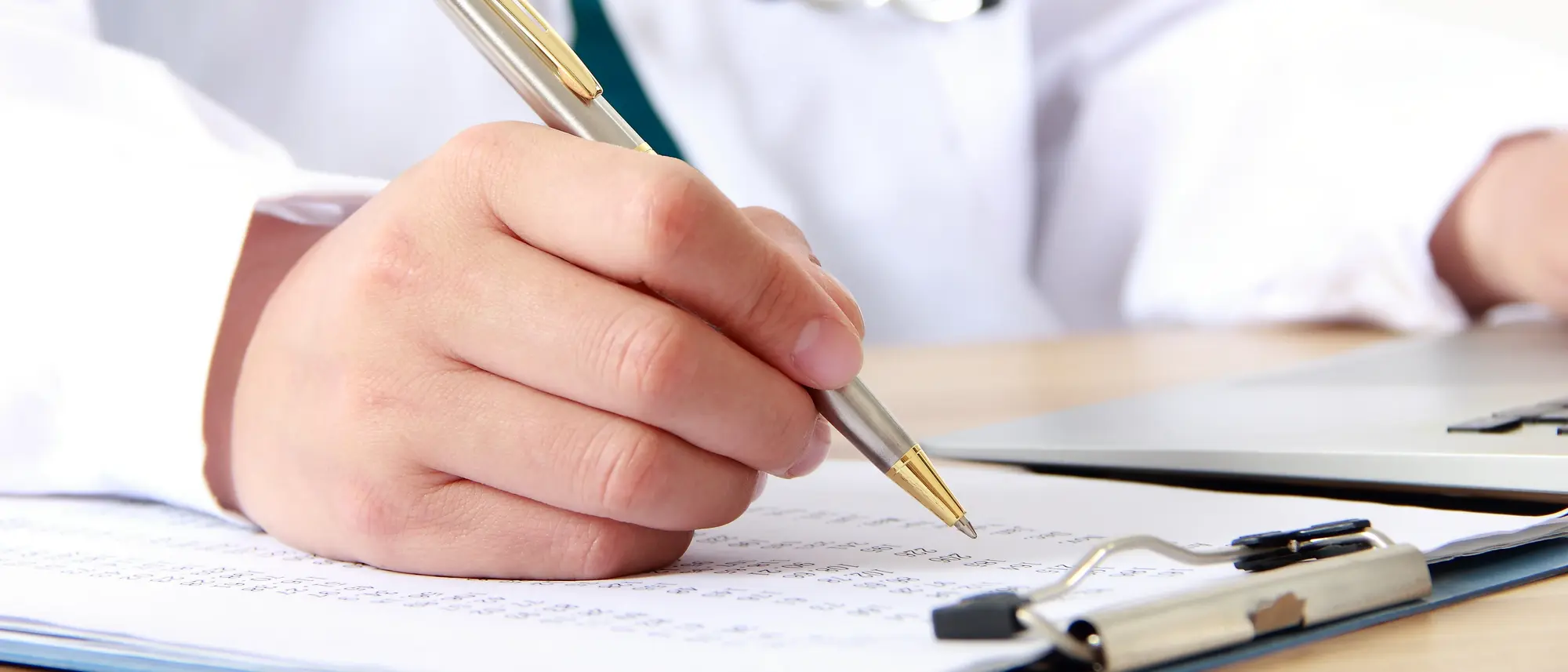 Eine Hand hält einen silbernen Stift zu sehen. Die Person schreibt etwas auf ein Blatt Papier, das auf einem Klemmbrett liegt. Der Hintergrund zeigt ein weißes Oberteil, das von einem Arzt oder einer Ärztin getragen wird, mit einem Stethoskop um den Hals.