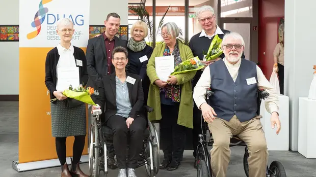 Gruppenfoto von sieben Personen während einer Veranstaltung der Deutschen Heredo-Ataxie-Gesellschaft (DHAG). Zwei Personen sitzen in Rollstühlen, fünf Personen stehen dahinter. Drei der stehenden Personen halten Blumensträuße und Urkunden in den Händen. Im Hintergrund ist ein Banner der DHAG mit dem Logo zu sehen.